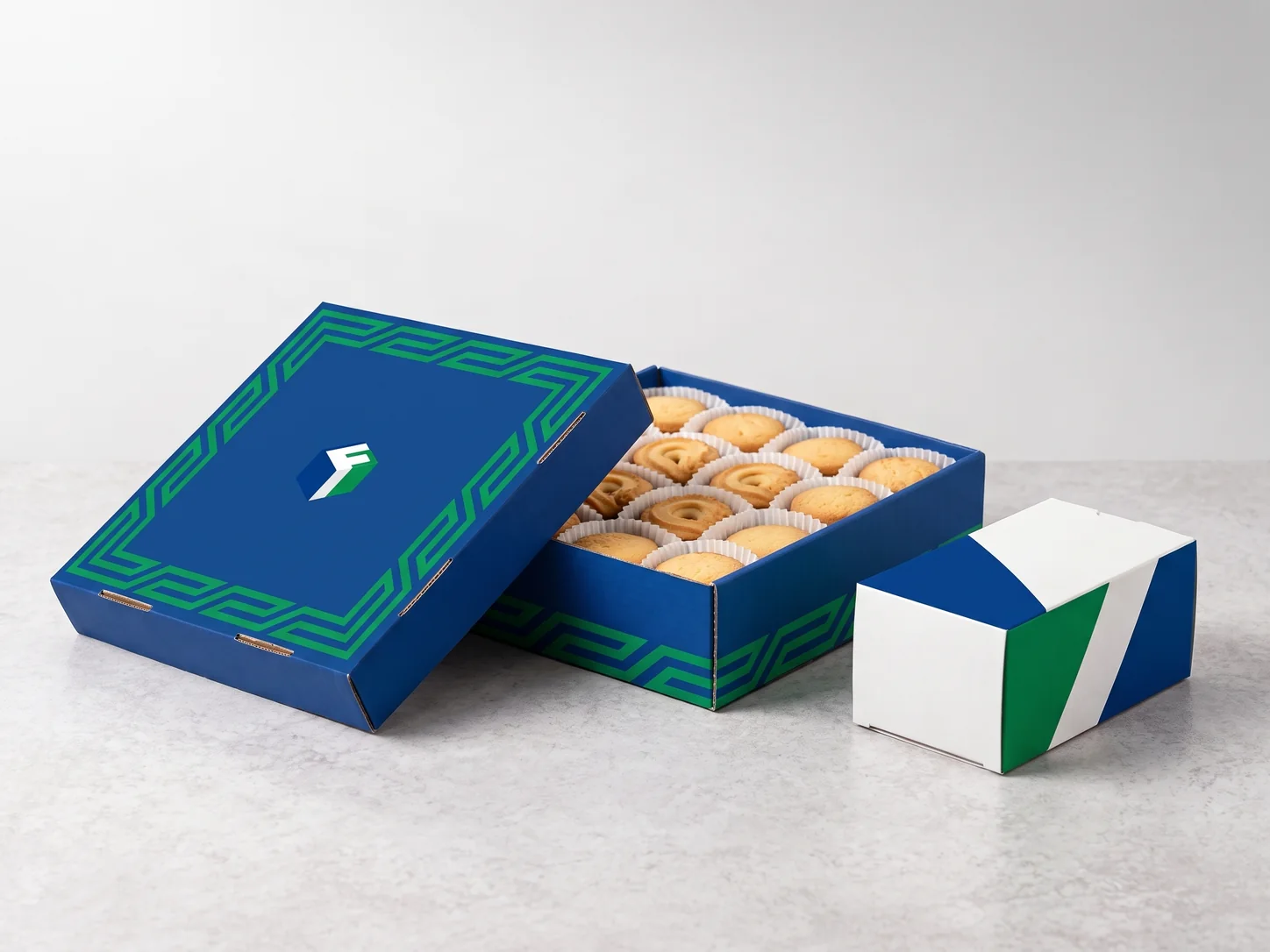 A blue printed biscuit gift box with its lid leaning beside it, full of golden butter biscuits in paper cups, next to a smaller retail carton.