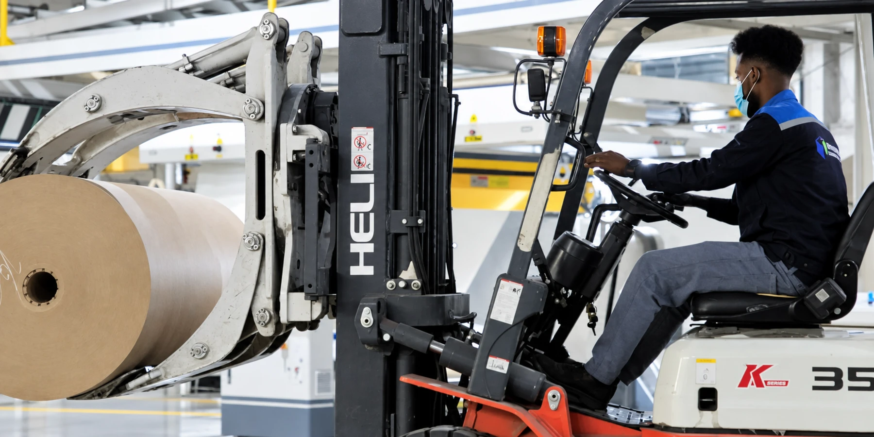 A forklift operator moves a large kraft paper reel beside the corrugator.