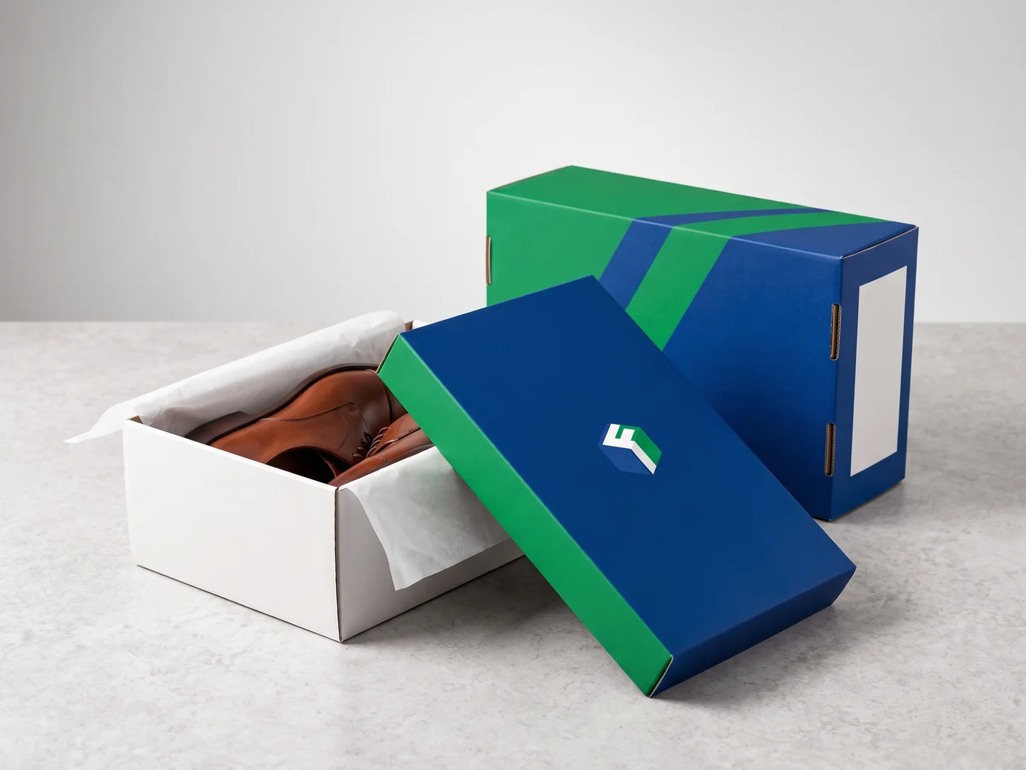 A white shoe box holding brown leather shoes on tissue paper, its blue and green lid leaning against it, with a second box behind.